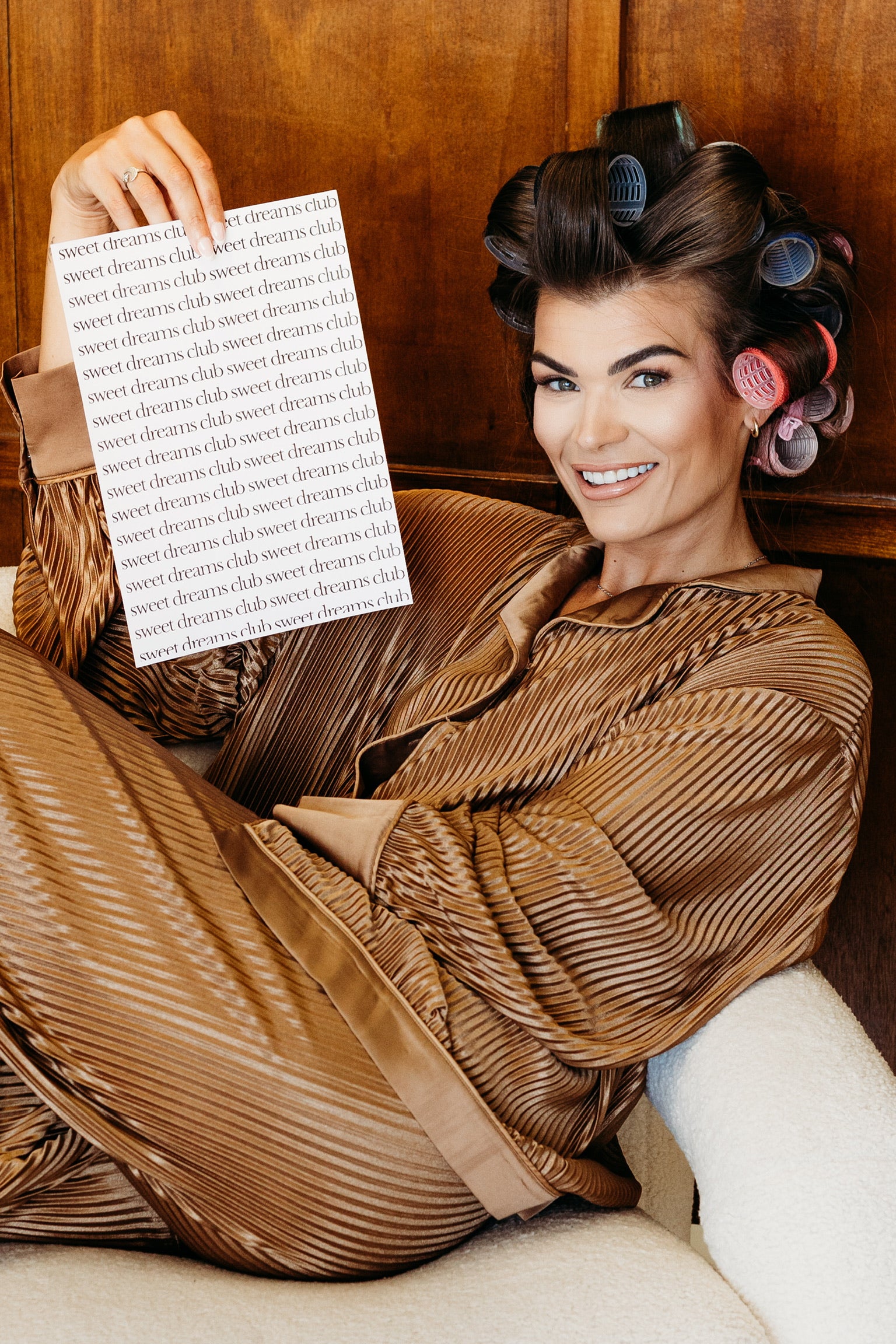 A woman reclining in a brown pleated robe with hair in curlers, holding up a sheet of paper covered in the repeating text 'sweet dreams club'. The background is a warm wood-paneled wall.