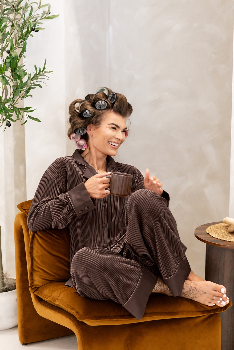 A woman wearing a chocolate brown pajama set sits on a mustard yellow armchair, holding a dark brown mug. She has her hair in curlers and is smiling in a cozy indoor setting with a potted olive tree and a small side table.