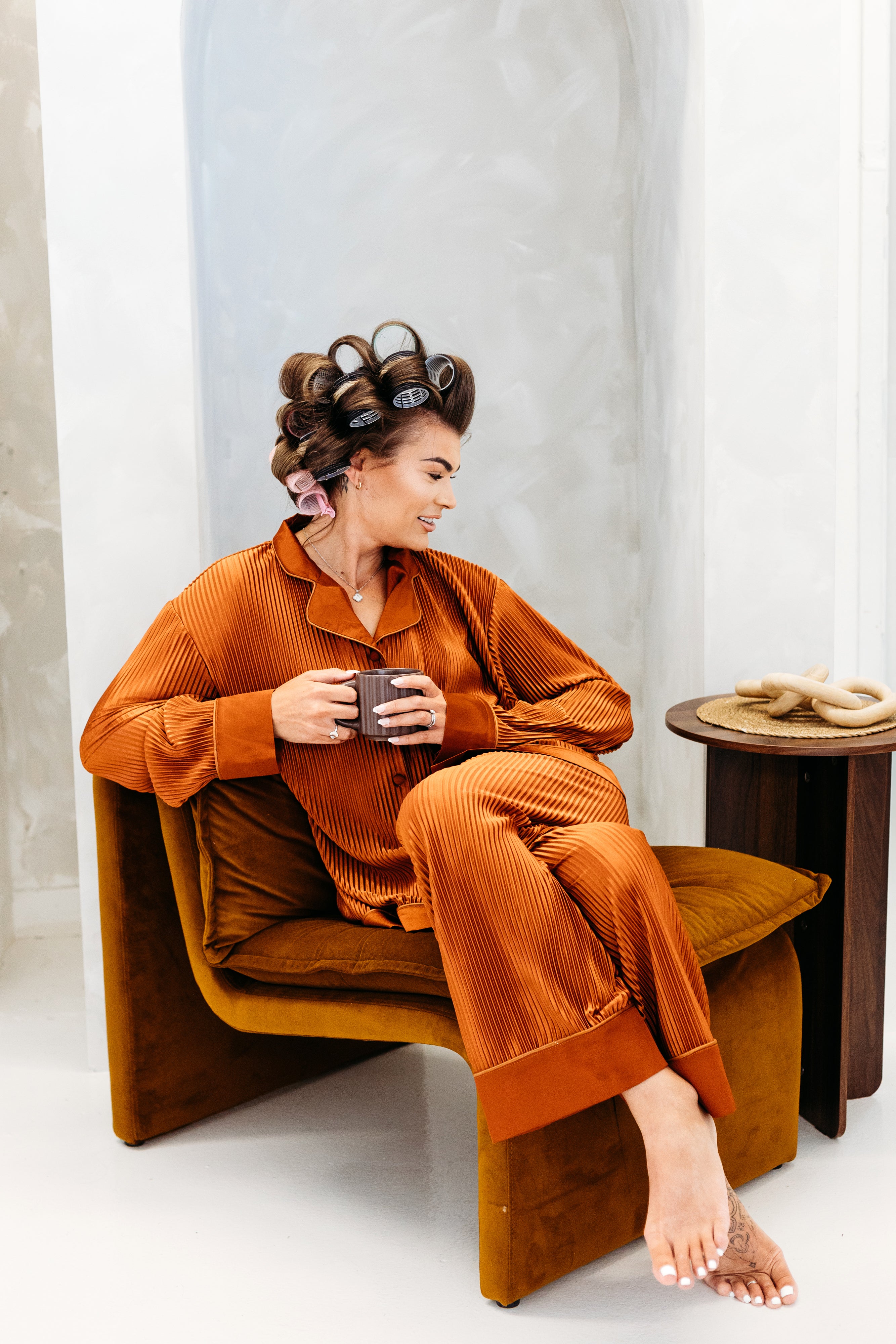 Woman in orange corduroy pajama set sitting on a brown chair holding a mug.