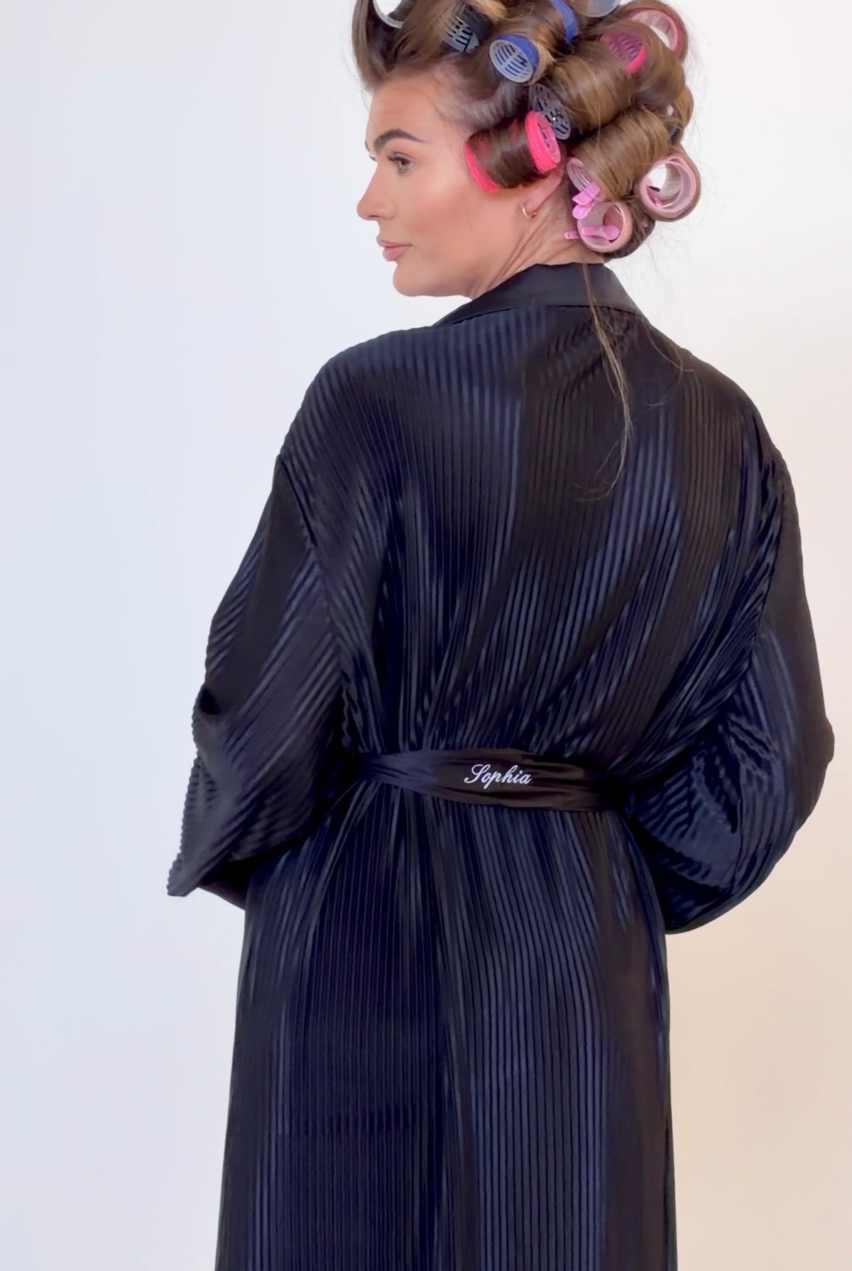 A woman shown from the back wearing a dark black plisse robe with a matching belt. Her hair is styled with large curlers in pink, purple, and blue, set against a plain light background.