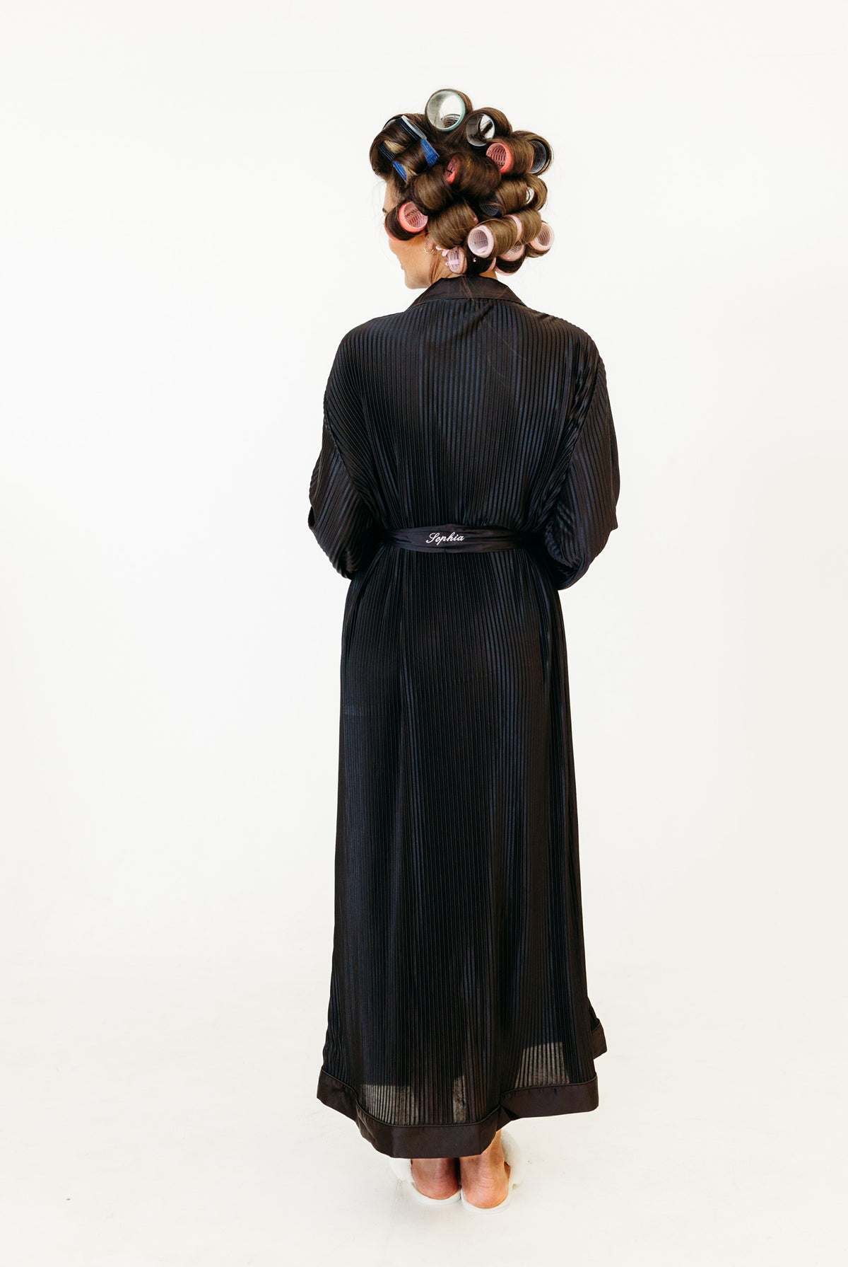A person shown from the back wearing a long black robe with subtle vertical pinstripes and a matching belt, styled with hair in curlers against a plain white background and a personalised belt
