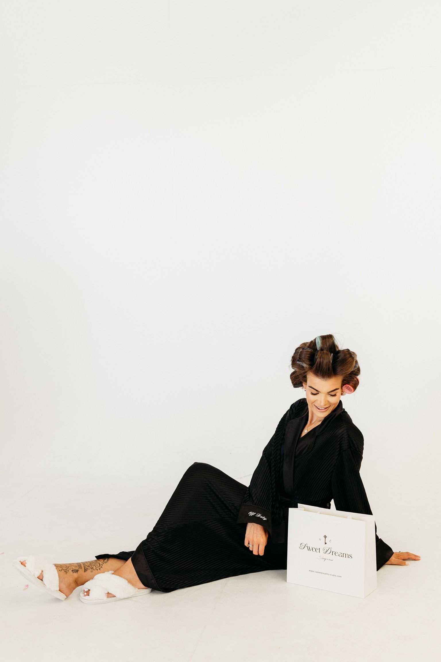A woman in a black plisse robe and white slipper sits on a white studio floor next to a white shopping bag printed with the brand name 'sweet dreams sleepwear'.