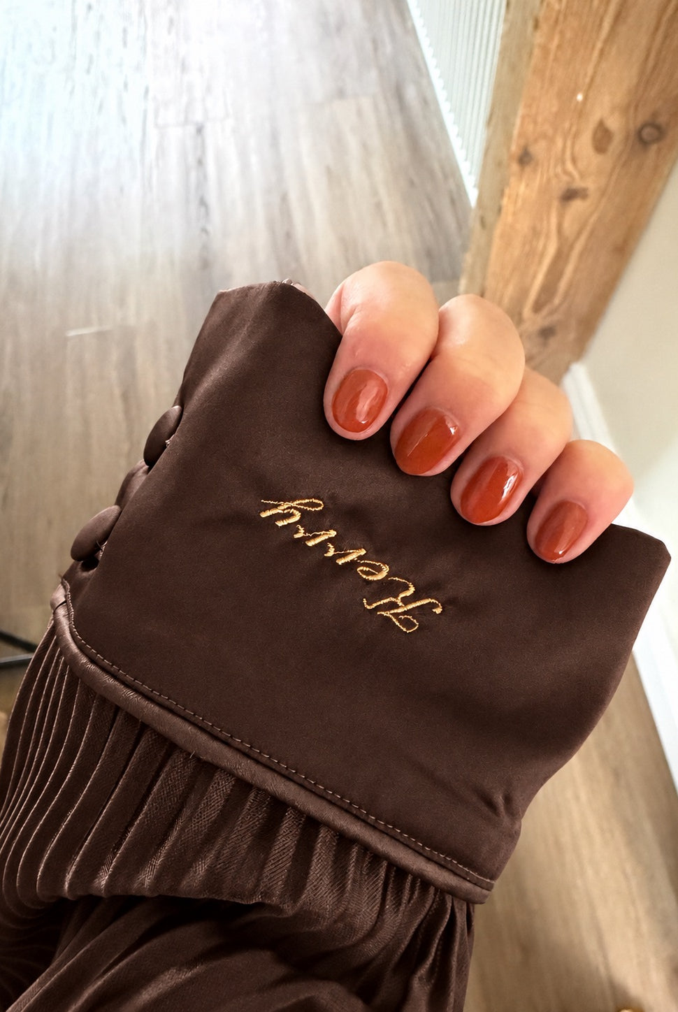 A close-up of a hand with orange-brown manicured nails holding a brown pjs cuff . The fabric has gold embroidery reading 'kerry' and a plisse brown texture along the lower edge.
