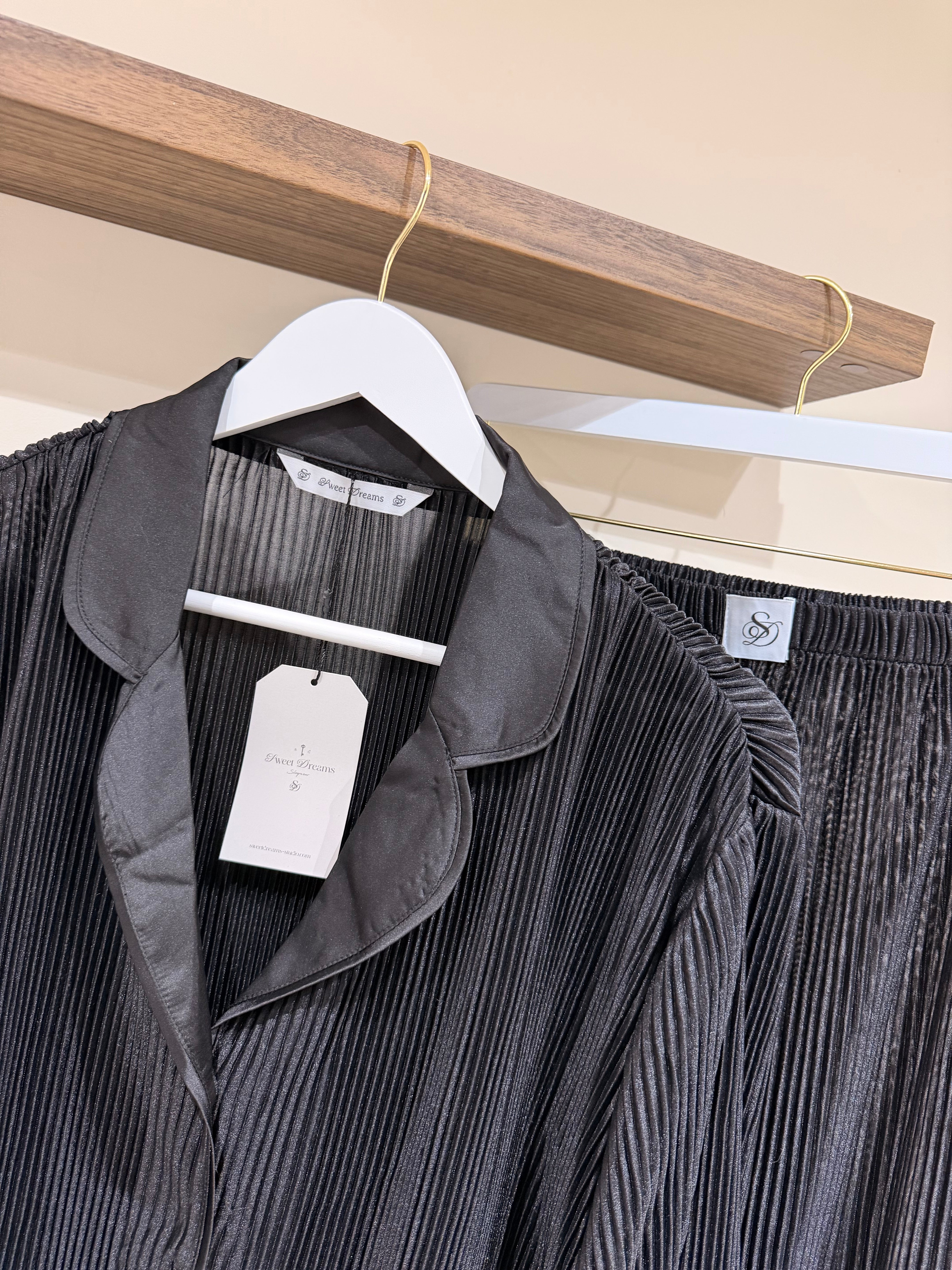 Black pleated pjs with tags on a wooden hanger against a neutral background