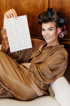 A woman reclining in a brown pleated robe with hair in curlers, holding up a sheet of paper covered in the repeating text 'sweet dreams club'. The background is a warm wood-paneled wall.