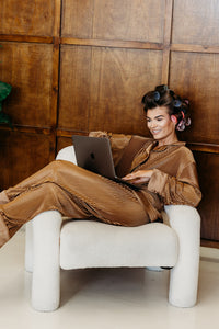 A woman reclining in a white armchair against a wood-paneled wall, wearing a matching brown ribbed top and trousers set and using a laptop.