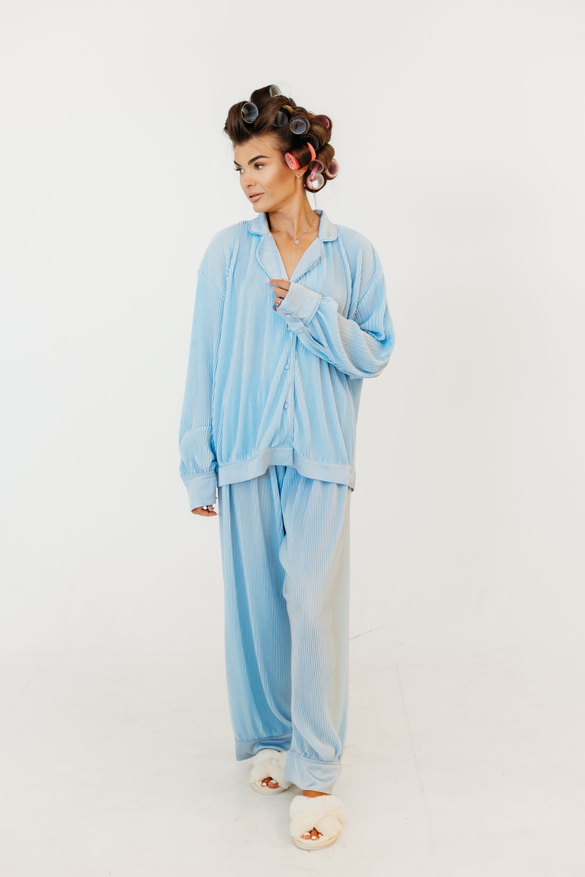 A woman modeling a light blue striped pajama set consisting of a long-sleeve button-up shirt and matching wide-leg pants, styled with white slippers and brown hair in curlers against a plain white studio background.