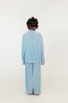A person viewed from behind wearing a light blue velour two-piece tracksuit with a long-sleeve top and wide-leg pants, styled with dark brown hair in curlers, against a plain white studio background.