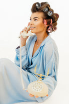 A woman in a light blue nightwear set sits on a white studio background, holding a cream rotary telephone receiver and looking upward. She has brown hair styled with pink and gray curlers.