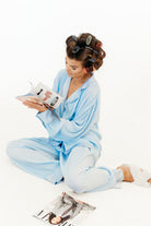 A woman sitting cross-legged on a white studio floor wearing a light blue pajama set, with hair rollers in her hair and reading a vogue magazine. Grazia magazine lies on the floor in front of her.
