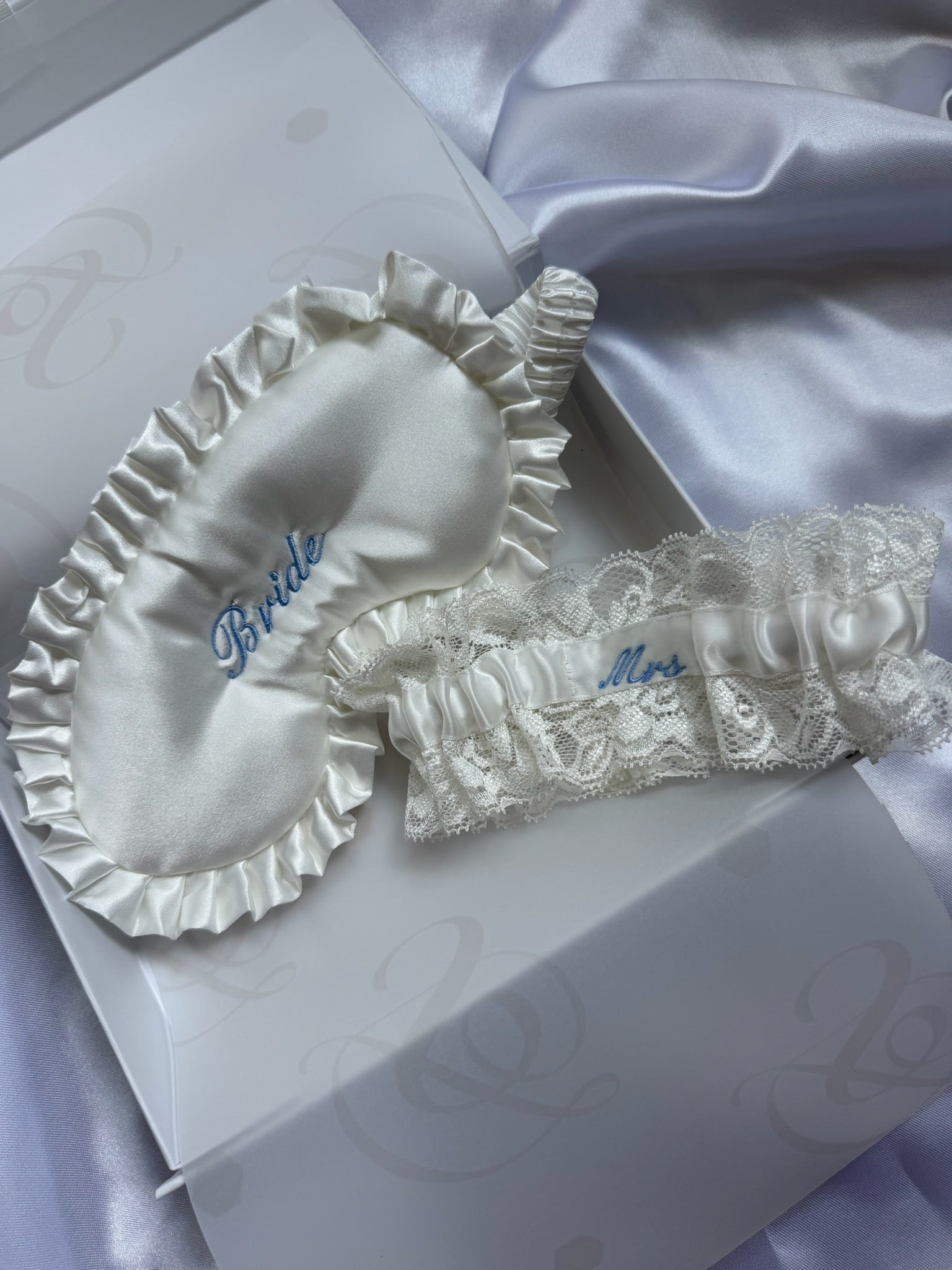 White bridal sleepwear set featuring a satin eye mask and lace garter belt with blue embroidered text, displayed on a white gift box with lavender satin fabric in the background.