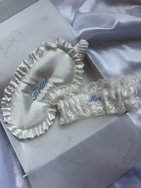 White bridal sleepwear set featuring a satin eye mask and lace garter belt with blue embroidered text, displayed on a white gift box with lavender satin fabric in the background.