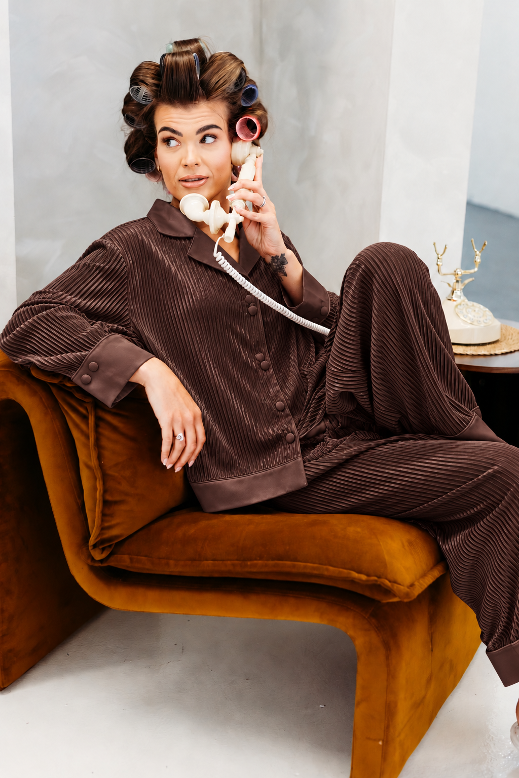 A woman wearing a brown  pajama set with matching pants and a button-up top sits on an orange-brown velvet lounge chair, holding a vintage cream rotary telephone receiver.