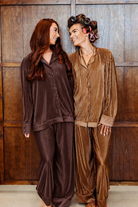 Two women wearing matching ribbed plisse  pajama sets, one in dark brown and one in tan, standing in front of a wooden wall.