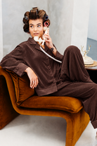A woman wearing a brown burgundy  pajama set with matching pants and a button-up top sits on an orange-brown velvet lounge chair, holding a vintage cream rotary telephone receiver.