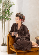A woman wearing a brown ribbed pajama set sits cross-legged on a mustard velvet chair, holding a mug. She has brown hair styled in curlers with pink and gray rollers.
