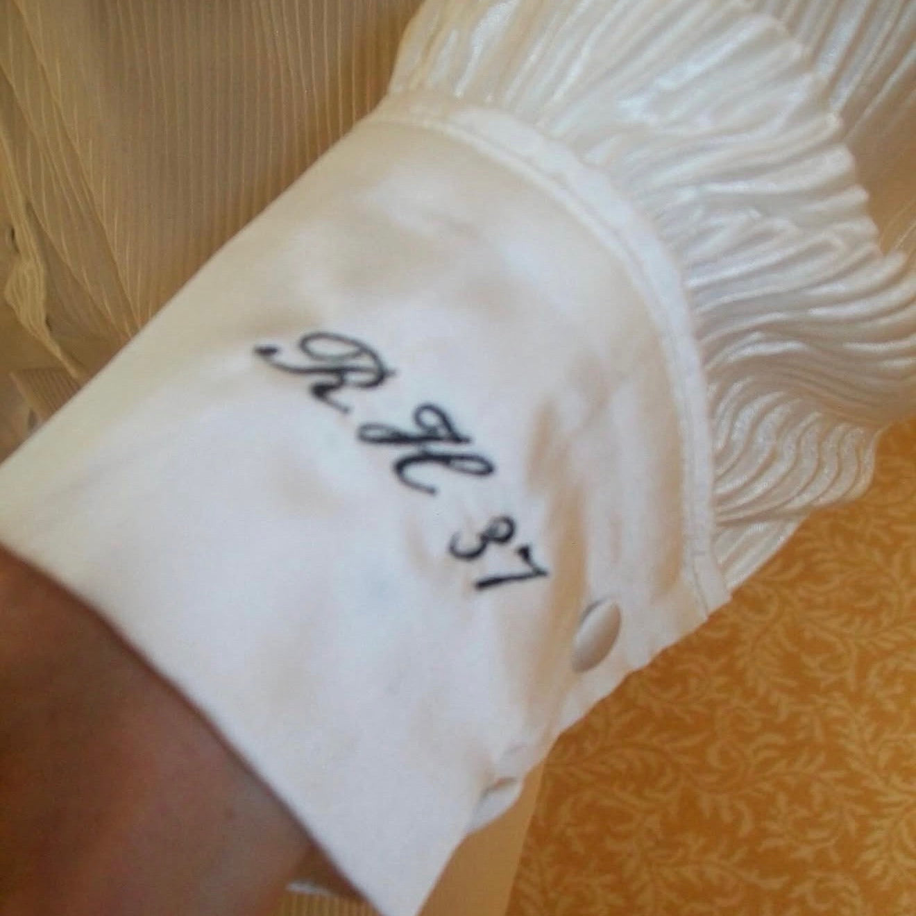 A close-up of a white ruffled sleeve or cuff with black embroidered script initials 'RH 31' on the front, worn on a person's wrist against a patterned gold fabric background.