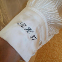 A close-up of a white ruffled sleeve or cuff with black embroidered script initials 'RH 31' on the front, worn on a person's wrist against a patterned gold fabric background.