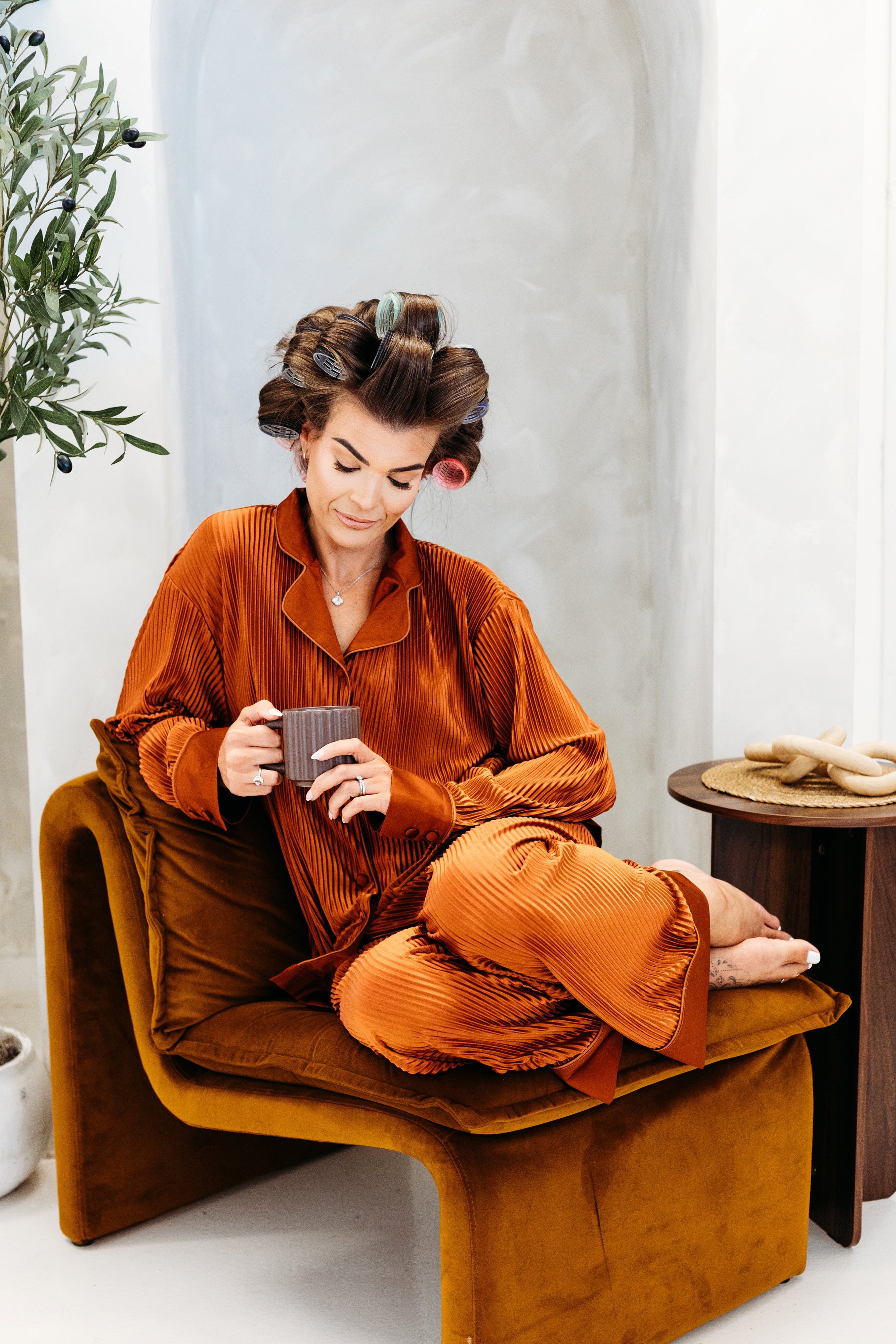 Woman in orange corduroy pajama set with hair in rollers, sitting on a brown chair indoors.