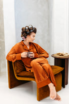 Woman in orange corduroy pajama set sitting on a brown chair holding a mug.