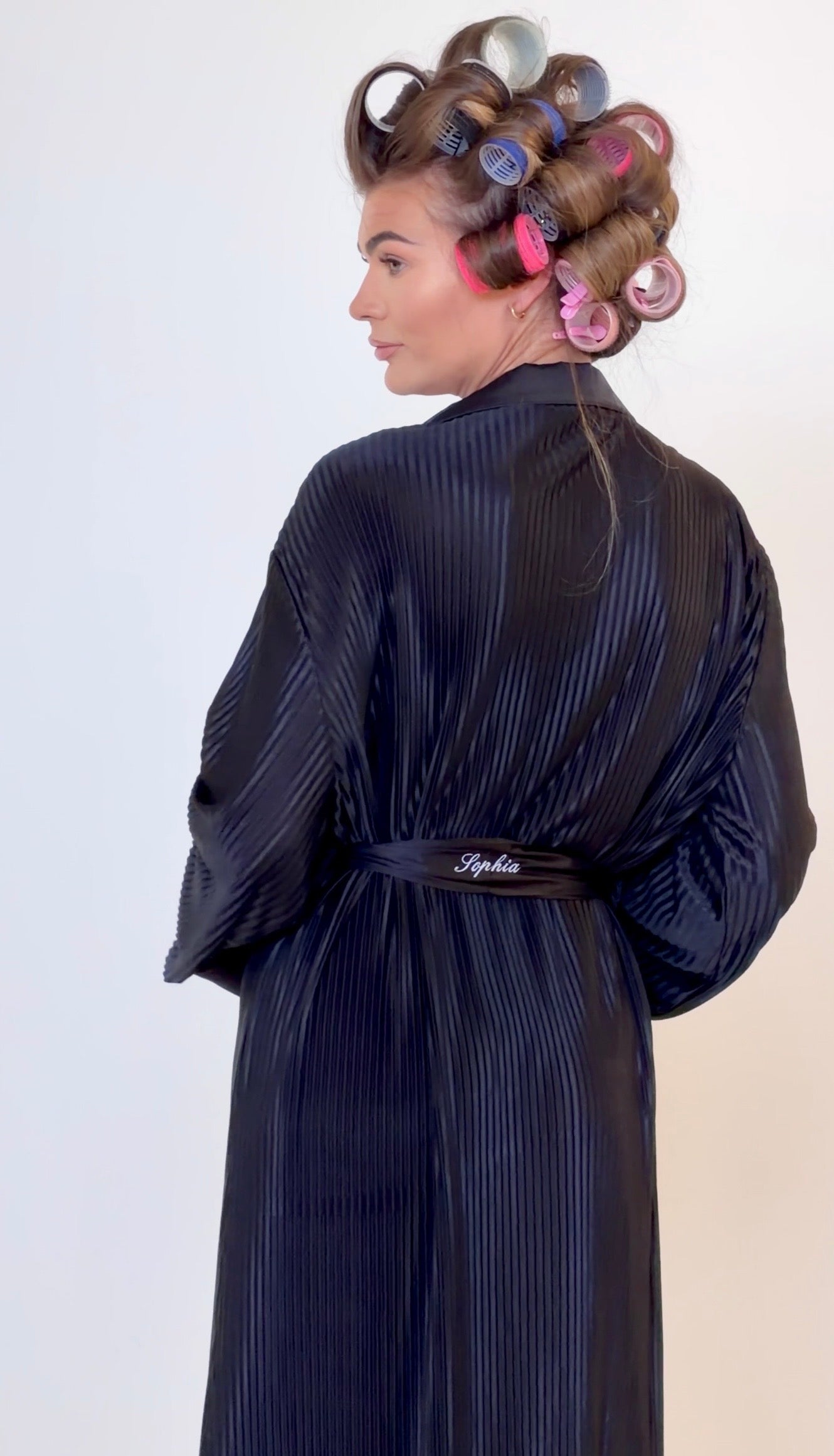 A woman shown from the back wearing a dark black plisse robe with a matching belt. Her hair is styled with large curlers in pink, purple, and blue, set against a plain light background.