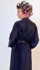 A woman shown from the back wearing a dark black plisse robe with a matching belt. Her hair is styled with large curlers in pink, purple, and blue, set against a plain light background.