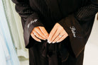 A close-up of a person wearing a black pleated robe with wide cuffs. The left cuff has the words 'Off Duty' embroidered in white script, and the right cuff has 'Ghster' embroidered in white script.
