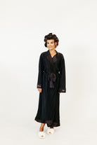 A woman wearing a long black silk-like robe with a tie waist and white slippers, standing against a plain white studio background.