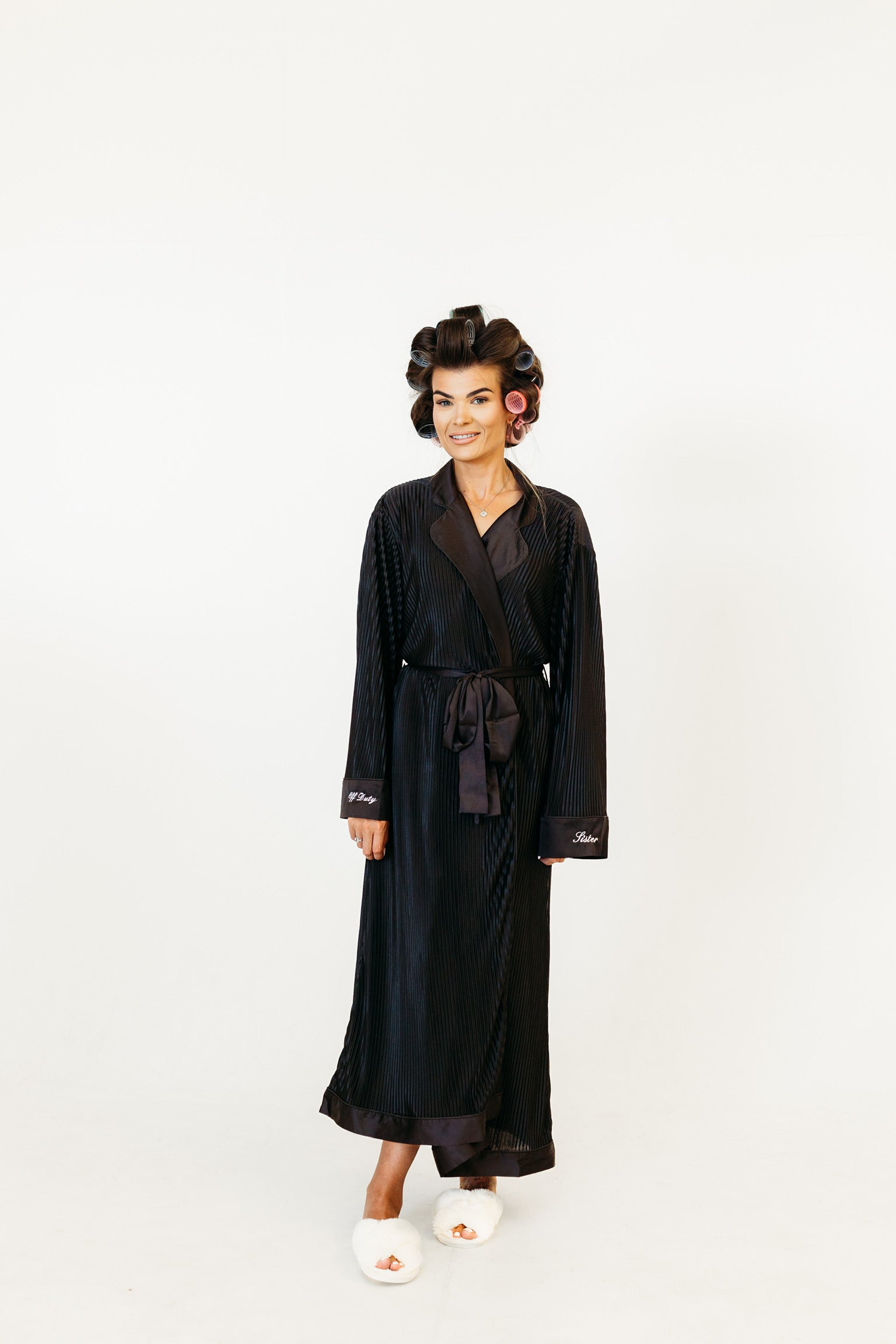 A woman wearing a long black silk-like robe with a tie waist and white slippers, standing against a plain white studio background.