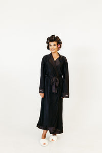 A woman wearing a long black silk-like robe with a tie waist and white slippers, standing against a plain white studio background.