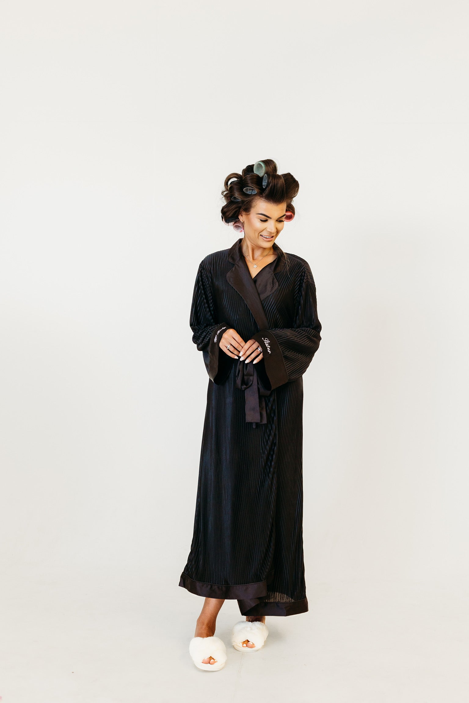 A woman wearing a long black satin robe with a shawl collar and tie belt, styled with vintage curlers in her hair and white slippers, standing against a plain light gray background.