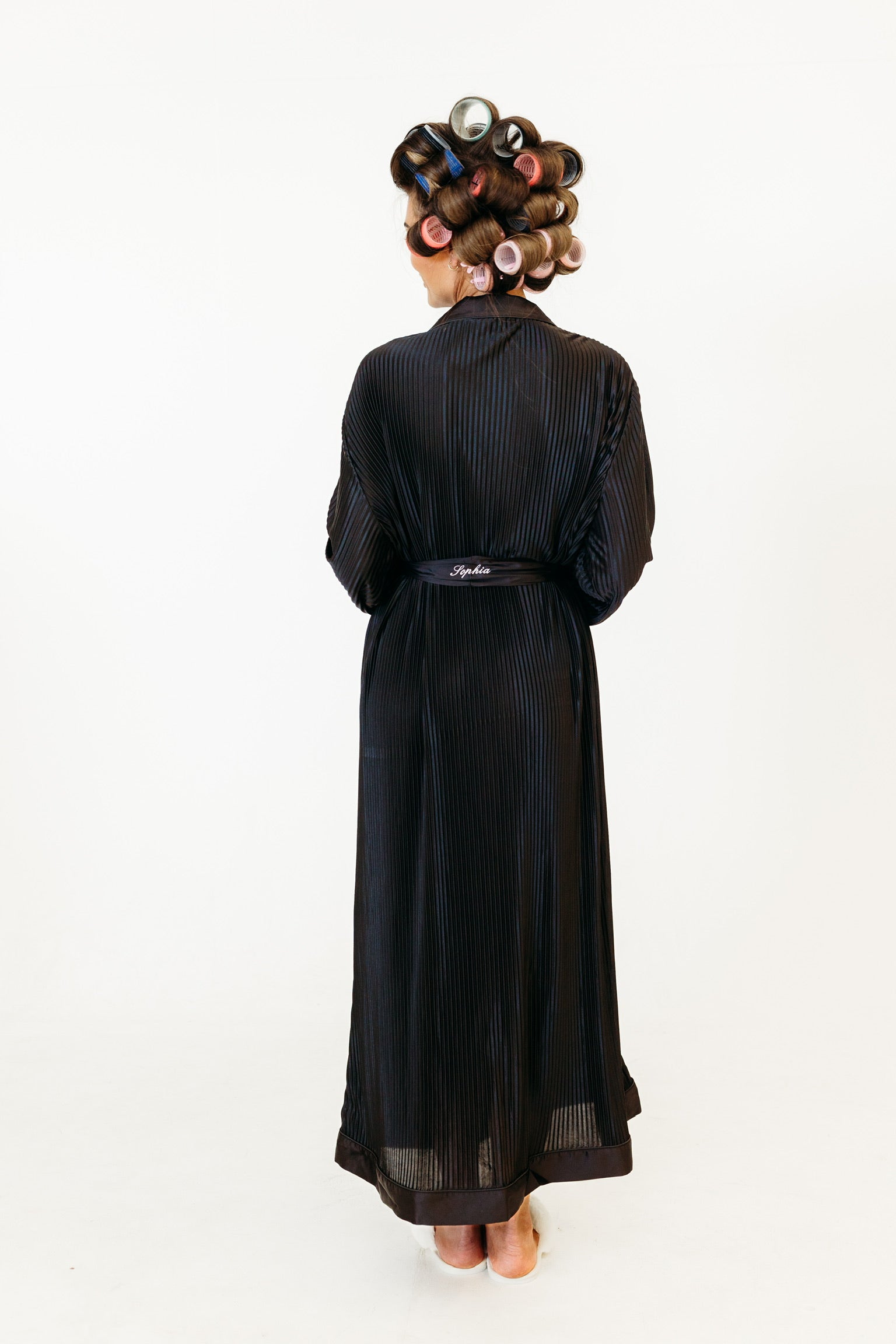 A person shown from the back wearing a long black robe with subtle vertical pinstripes and a matching belt, styled with hair in curlers against a plain white background.