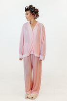 A woman modeling a light pink two-piece pajama set consisting of a long-sleeve button-front top and matching wide-leg pants, photographed against a plain white studio background.