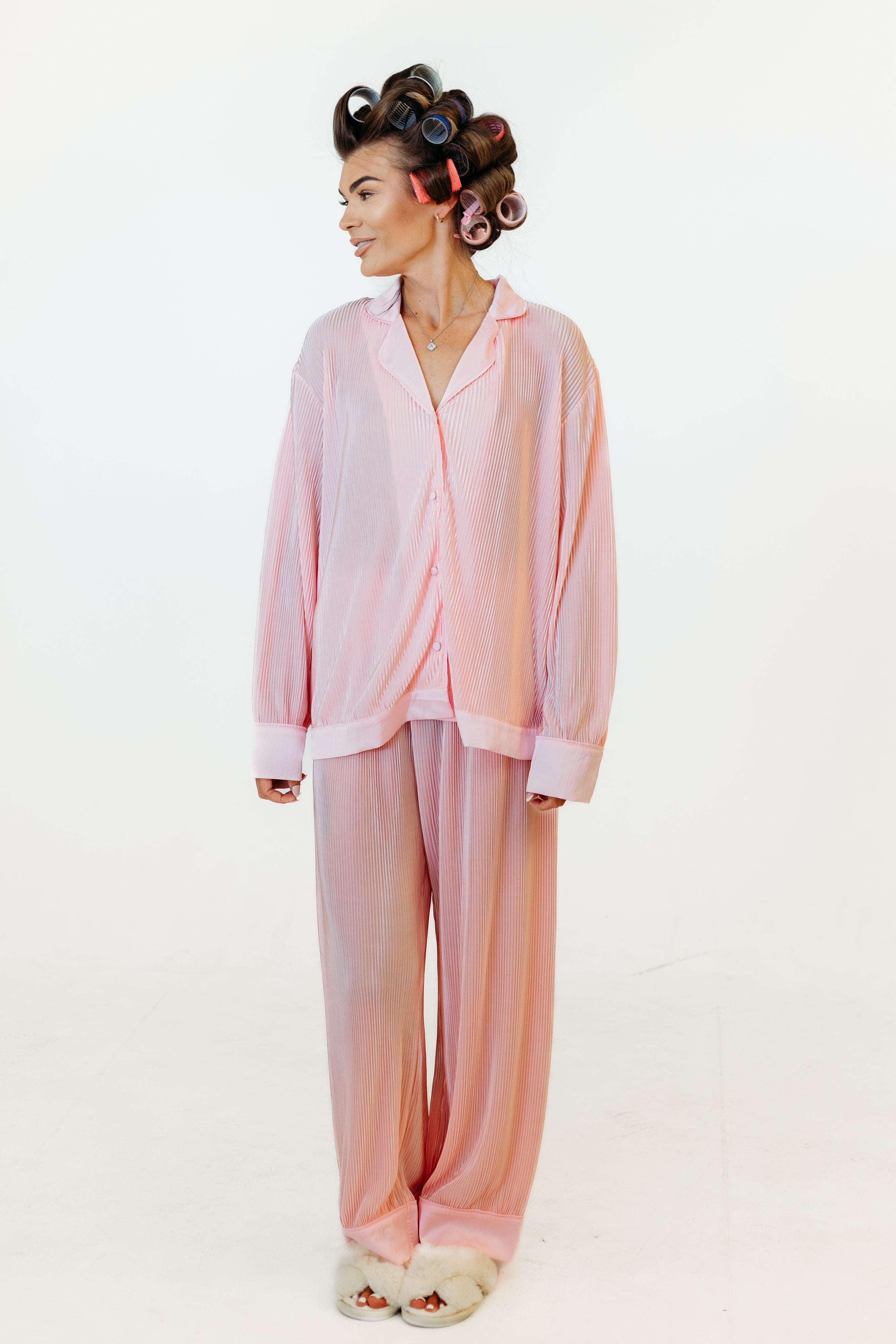 A woman modeling a light pink two-piece pajama set consisting of a long-sleeve button-front top and matching wide-leg pants, photographed against a plain white studio background.
