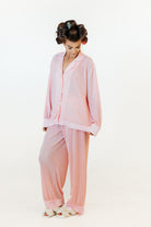 A woman wearing a light pink two-piece pajama set with a long-sleeve button-front top and matching wide-leg pants, standing against a plain white studio background.