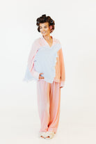 A woman modeling a pastel sleepwear set consisting of a light pink long-sleeve top and matching wide-leg pants, styled with a light blue shawl and white slippers against a plain white background.