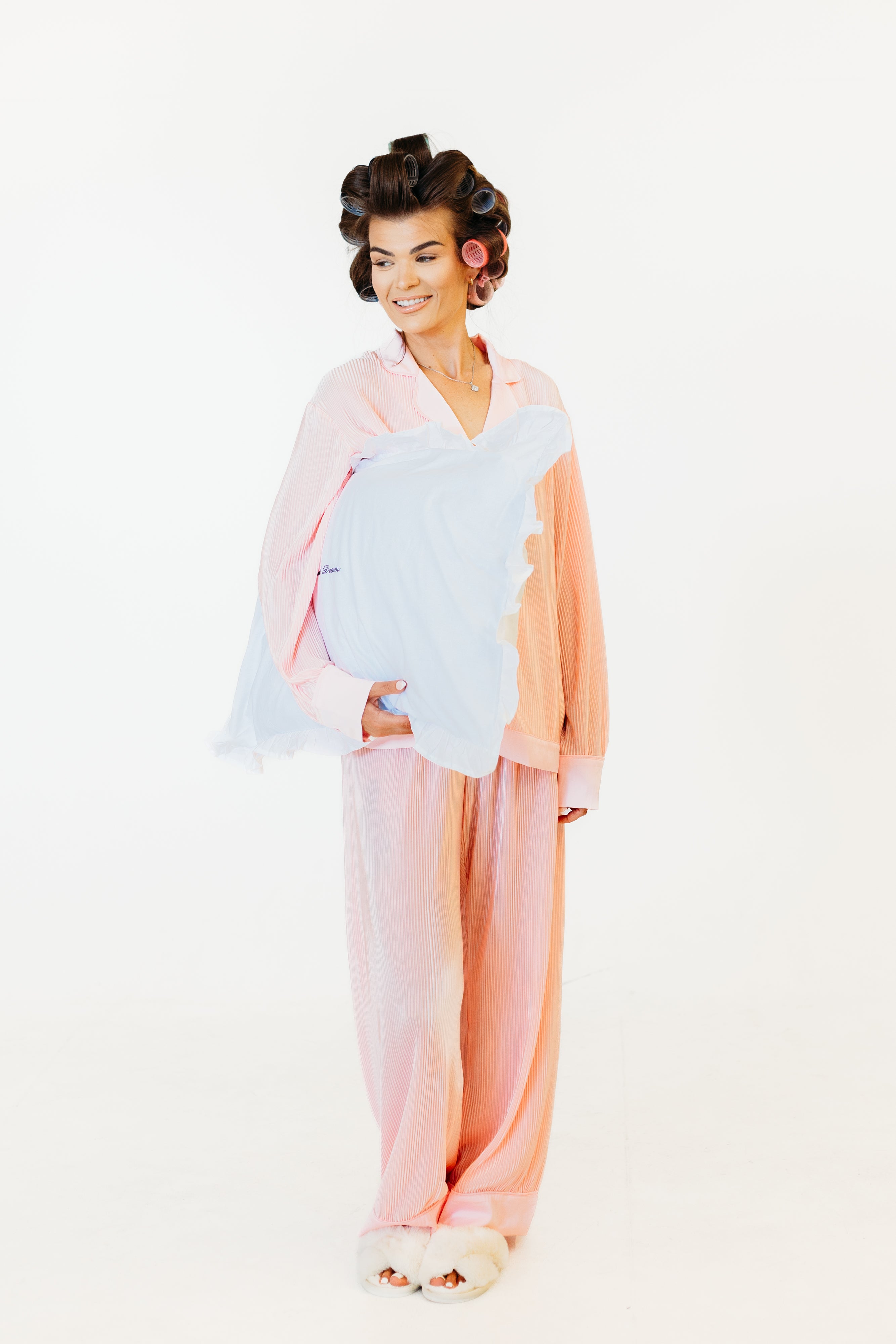 A woman modeling a pastel sleepwear set consisting of a light pink long-sleeve top and matching wide-leg pants, styled with a light blue shawl and white slippers against a plain white background.