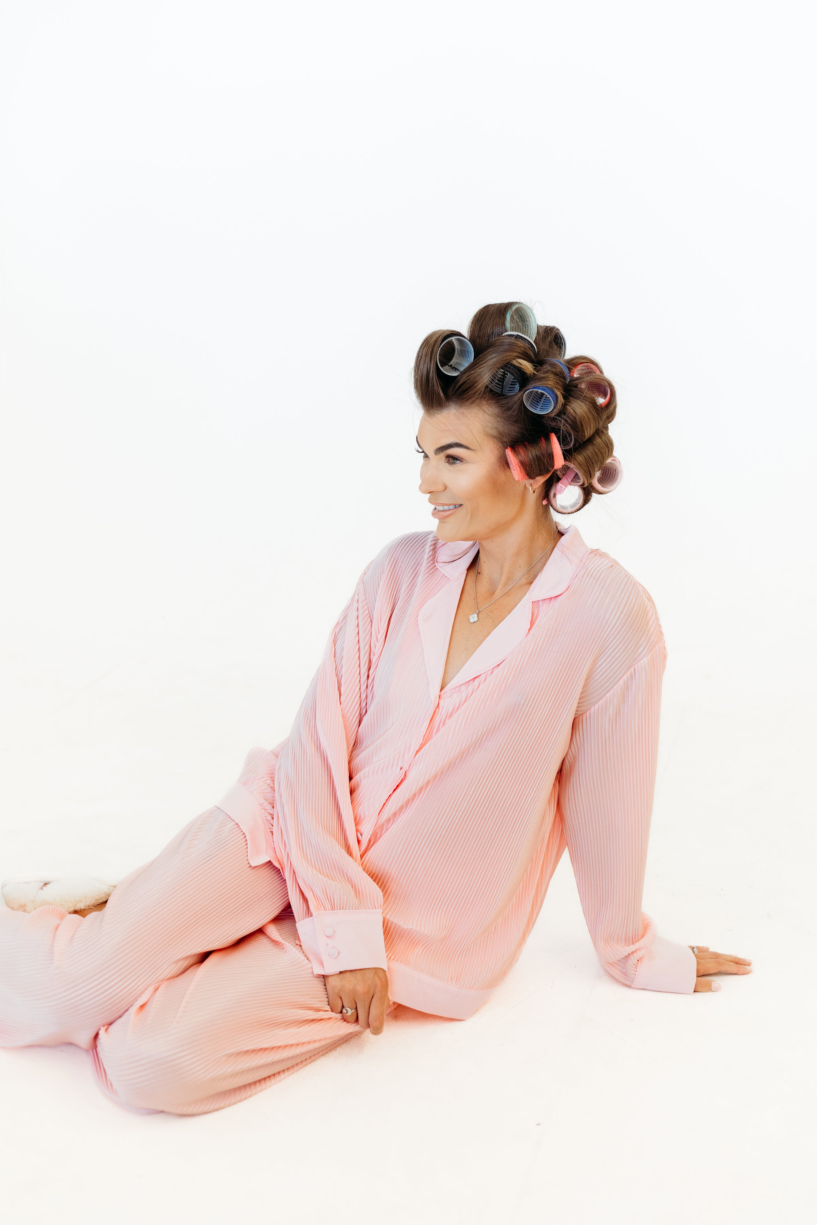 A woman wearing a light pink pajama set sits on a white studio floor with her hair in dark pink curlers.