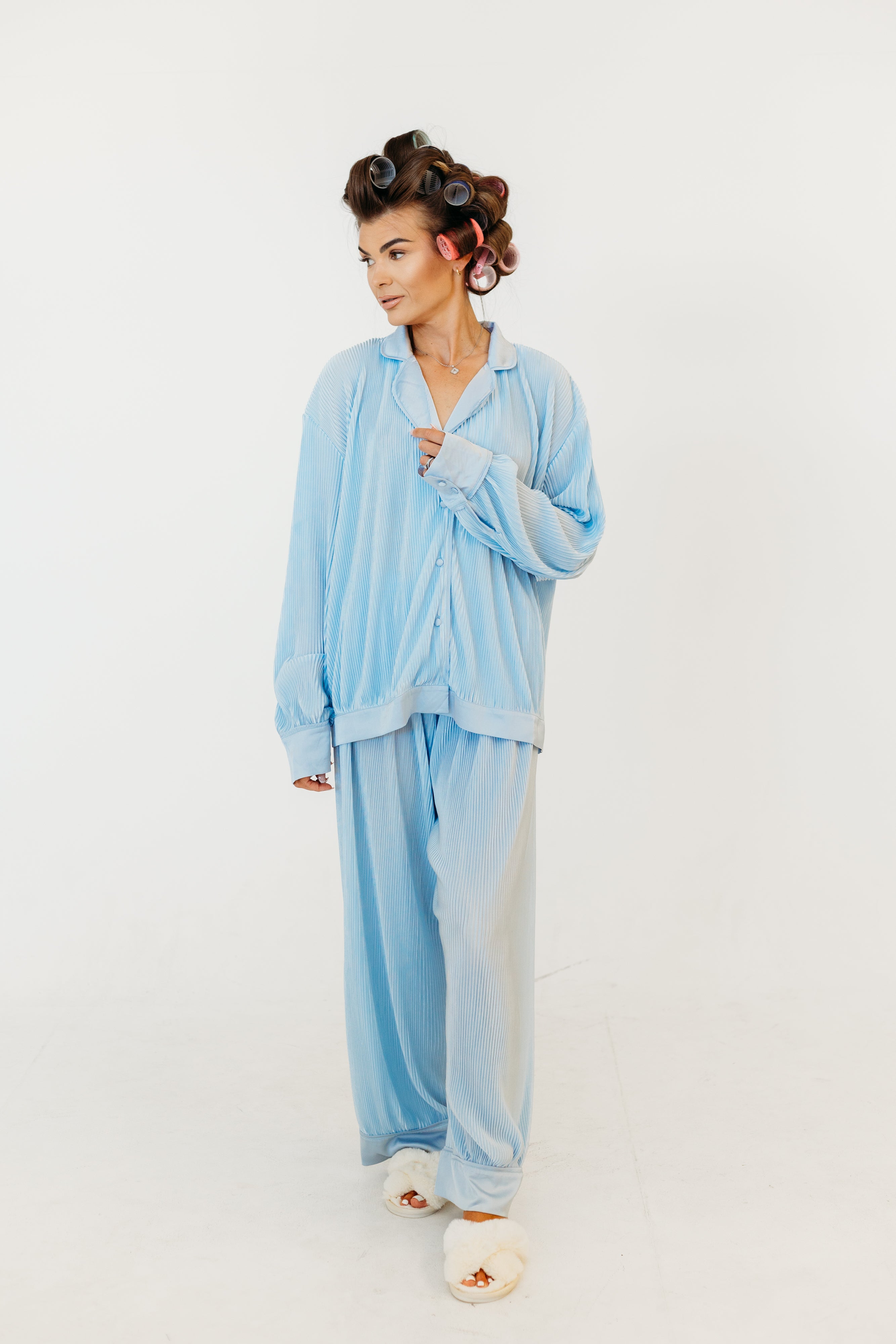 A woman modeling a light blue striped pajama set consisting of a long-sleeve button-up shirt and matching wide-leg pants, styled with white slippers and brown hair in curlers against a plain white studio background.