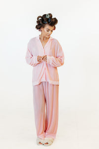 A woman wearing a light pink pajama set with a long-sleeve button-front top and matching pants, standing against a plain white studio background. She has dark hair styled in curlers and is wearing white slippers.