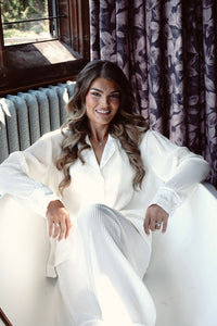 A woman with long wavy brown hair sits smiling in a white ribbed lounge set, featuring a matching long-sleeve collared top and wide-leg trousers. She is posed in a sunlit room with a window, a blue-gray radiator, and dark purple patterned curtains.