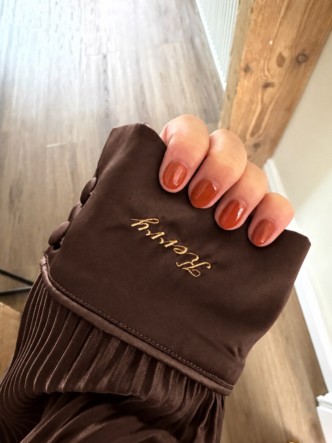 A close-up of a hand with orange-brown manicured nails holding a brown pjs cuff . The fabric has gold  embroidery reading 'kerry' and a plisse  brown texture along the lower edge.
