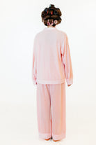 A person shown from the back wearing a light pink two-piece lounge set with a long-sleeve top and wide-leg pants, standing against a plain white studio background. The person has dark hair styled with curlers and pink hair accessories.