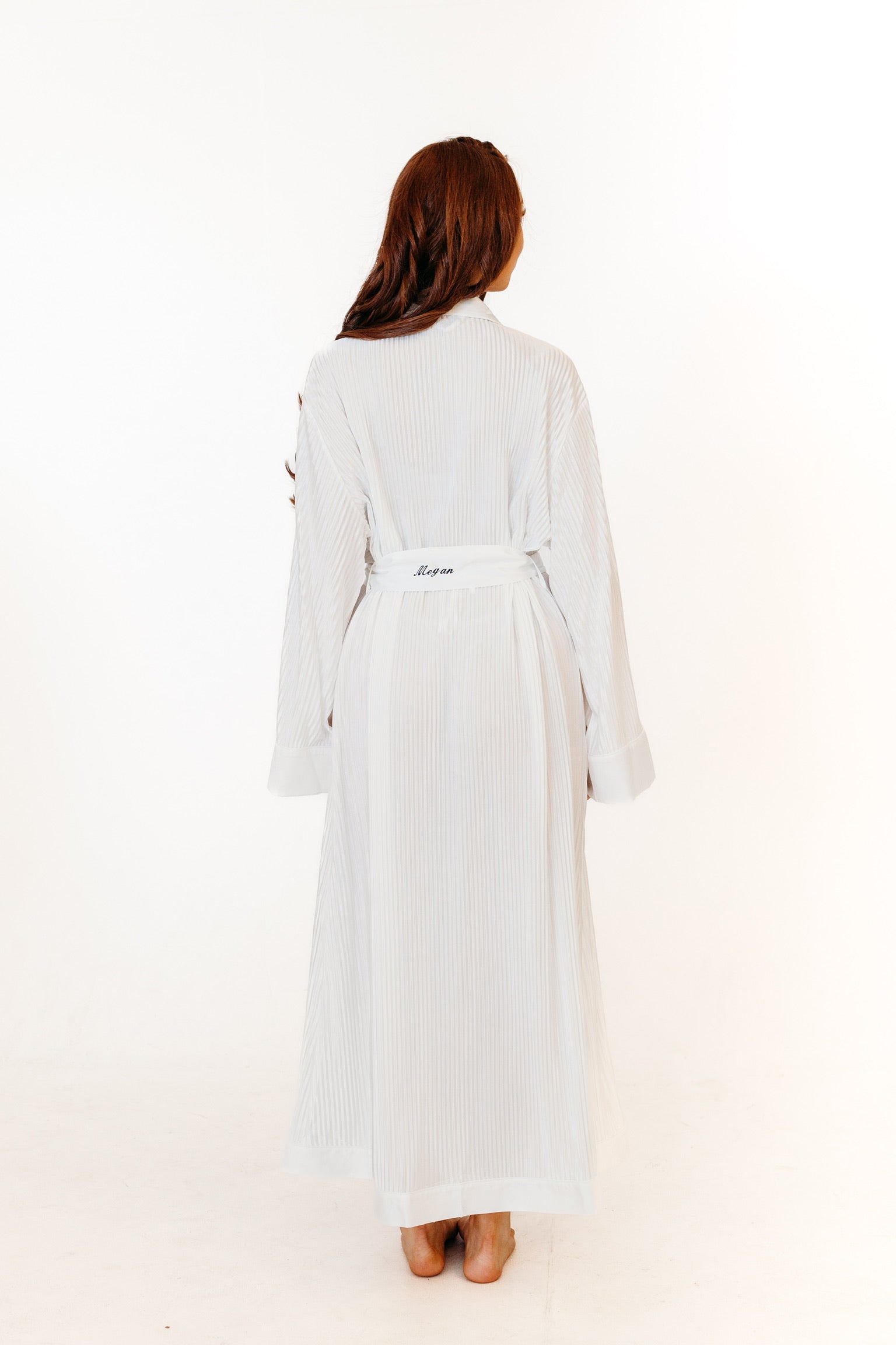 A woman viewed from behind wearing a long white robe with a tie belt, and personalisation standing against a plain white studio background.