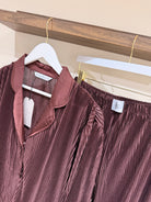 Burgundy  pleated shirt and pants on pyjamas set with tags against a wooden shelf.