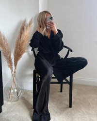 A person sitting on a chair wearing a black pyjama with satin lapels and cuffs, featuring a relaxed fit.